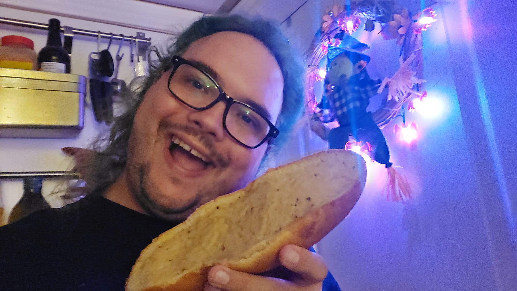 A man with long green hair and no beard holds a piece of garlic bread in a kitchen lit in purple light, Photo 3